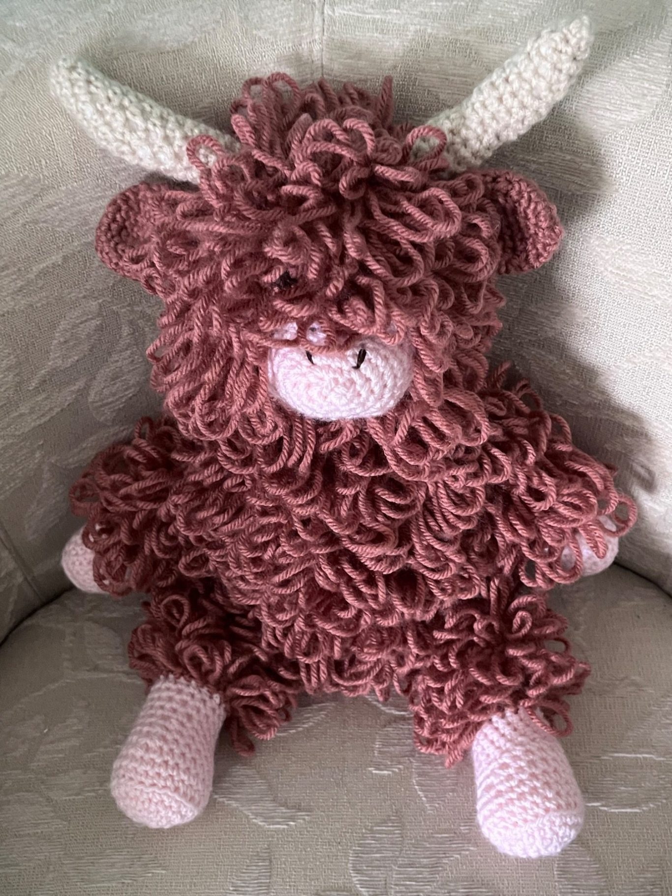 A fluffy, russet and  pink crochet toy cow with curly fur and horns.